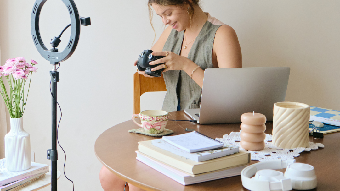 Woman sitting at a table with a laptop, camera, and lighting equipment in a home setting.